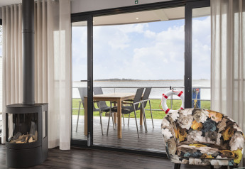 Salón de casa flotante con chimenea, silla floral y terraza con vistas al agua en Friesland, Países Bajos.