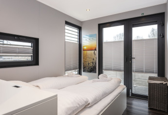 Moderne slaapkamer op een woonboot met tweepersoonsbed, grote ramen en zonsondergang schilderij aan de muur.