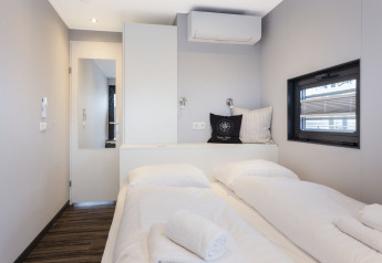 Moderne, lichte kamer op woonboot met tweepersoonsbed, raam, airco en handdoeken op bed in Friesland.