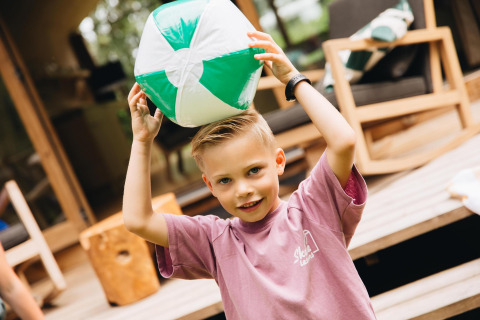 Jongen in roze T-shirt speelt met een groen-witte strandbal voor een lodge met houten tuinmeubelen.