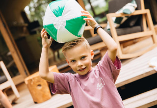 Jongen in roze T-shirt speelt met een groen-witte strandbal voor een lodge met houten tuinmeubelen.