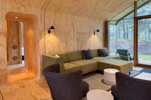 Moderne lodge woonkamer met houtpanelen, groene bank en grote vensters in Wikkelhouse Duo, Beerze Bulten.
