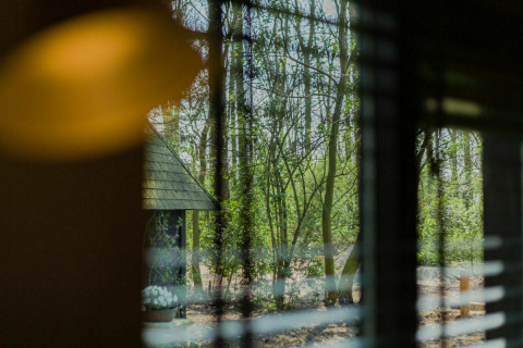 Udsigt gennem et vindue med persienner til skov og et lille hus ved Buitengoed Ruysbos, Nederlandene.