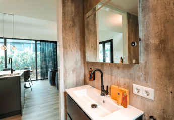 Interior de una cabaña moderna con lavabo, espejo, grifería elegante, cocina y grandes ventanas al jardín.