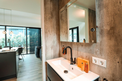 Interno di un lodge moderno con lavabo, specchio e rubinetto, vista su cucina open space e ampie finestre.