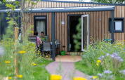 Dos personas sentadas fuera de una tiny house en Holiday Park 't Rheezerwold, Países Bajos, rodeados de flores silvestres.