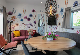 Salón moderno en una tiny house con pared floral, mesa redonda de madera y sofá junto a la ventana.