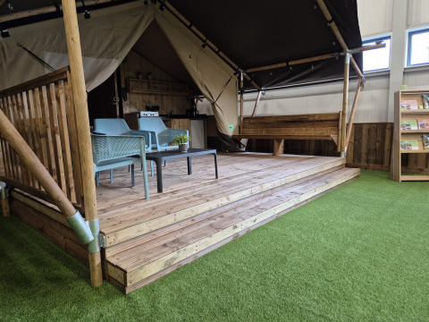 Tente safari avec sanitaires, terrasse en bois, coin salon et présentation de gazon artificiel intérieur.