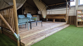 Tenda safari con servizi igienici, terrazza in legno, area salotto e erba artificiale all’interno.