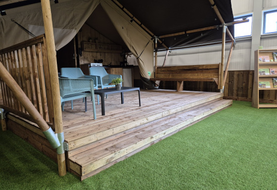 Safaritent met sanitaire voorzieningen, houten terras, zitgedeelte en kunstgras in een showroom.