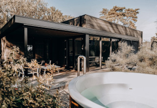 Lodge moderno con ventanales, terraza de madera y jacuzzi exterior rodeado de plantas y árboles.