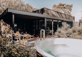 Lodge moderne avec grandes baies vitrées, terrasse en bois et jacuzzi extérieur entouré de verdure.