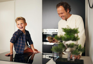Een vader en zijn zoon lachen samen in een moderne keuken, dicht bij een bonsaiboom en koffiekopje.
