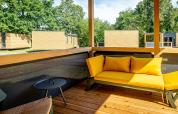 Terrazza in legno con divano giallo e tiny house moderne sullo sfondo a Urban-Gardens Lodge in Belgio.