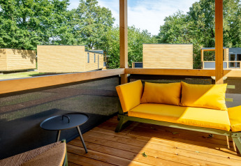 Wooden terrace with a yellow sofa and modern tiny houses in the background at Urban-Gardens Lodge, Belgium.