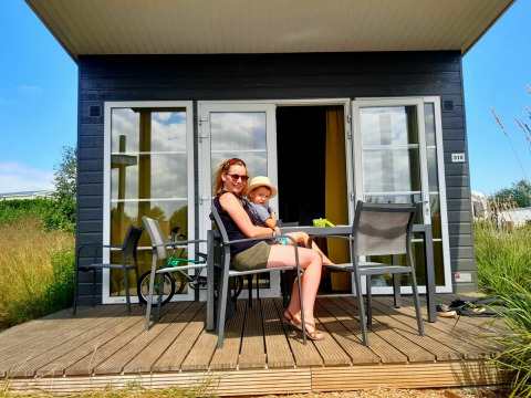 A woman and child sit on the wooden terrace in front of a Comfort Lodge tiny house at Kompas Camping Westende.