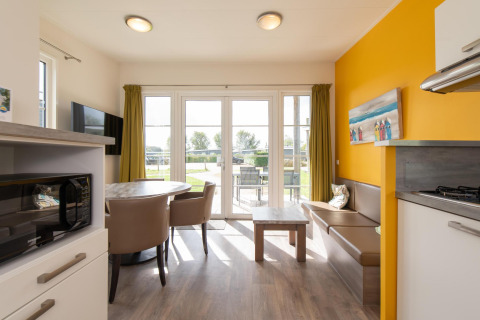 Helder glampingverblijf met eettafel, zitbank, kitchenette, gele muur en tuinzicht.