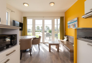 Helder glampingverblijf met eettafel, zitbank, kitchenette, gele muur en tuinzicht.
