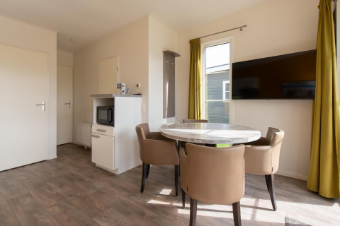 Moderne lodge bij Kompas Camping Westende met eettafel, vier stoelen, tv, magnetron en gele gordijnen.
