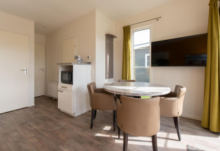 Moderne lodge bij Kompas Camping Westende met eettafel, vier stoelen, tv, magnetron en gele gordijnen.