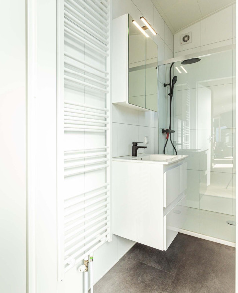 Baño moderno en Silva Wellness con tocador blanco, espejo grande, ducha negra y radiador para toallas.