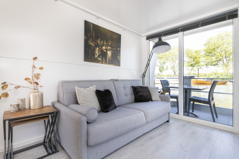 Lichte leefruimte op woonboot Harbour Lodge Amsterdam met zetel, decoratie en uitzicht op water.
