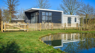 Moderne boslodge met afgesloten tuin en vijver, omringd door bomen en een stralende hemel.