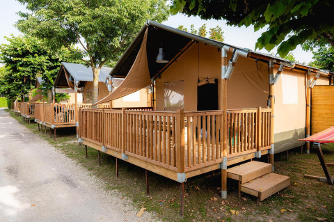 Tienda safari Villatent Outback con terraza de madera en Campeggio del Garda, Italia, rodeada de árboles verdes.