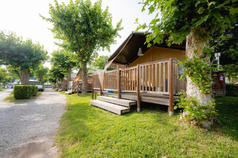 Tienda safari Villatent Outback con terraza de madera en Campeggio del Garda, Italia, rodeado de árboles verdes.