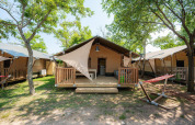 Safari-Zelt Villatent Outback mit Holzveranda, Hängematte und Bäumen auf sonnigem Campingplatz.