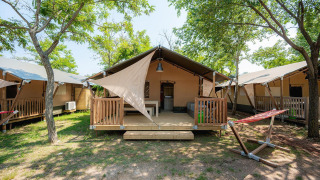Tente safari Villatent Outback avec véranda en bois, hamac et arbres sur un terrain de camping ensoleillé.
