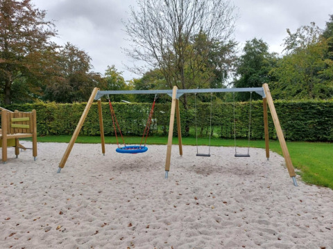 Parque infantil con columpios y arena, rodeado de setos en Camping de Beukenhof, Drenthe, Países Bajos.